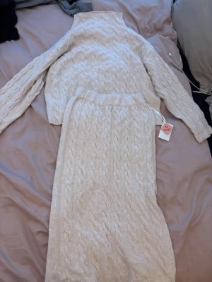 Beige kabelstickat set från ADIAZ - Säljer ett beige kabelstickat set från ADIAZ med långärmad tröja och matchande kjol. Setet har en mysig och trendig look med bred resår i midjan på kjolen och hög hals på tröjan. Perfekt för dig som gillar stickade plagg med snygg struktur.
