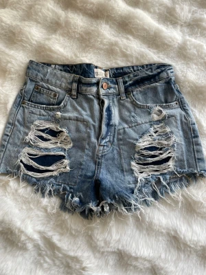 Blå slitna jeansshorts med fransar - Säljer ett par blå denimshorts med råa kanter och stora slitningar framtill. Shortsen har fem fickor, bälteshällor och knappgylf. Perfekta för sommaren och ger en avslappnad, trendig look.