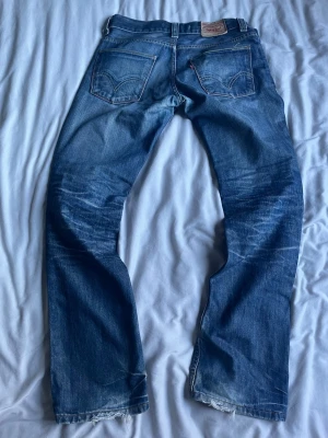 Blå Levi's slimfit jeans - Klassiska blå jeans från Levi's med rak passform och slitna detaljer vid bensluten. Jeansen har fem fickor, synliga sömmar och en ikonisk läderpatch bak i midjan. Perfekta för en avslappnad och tidlös stil.