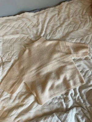 Beige stickad tröja med hög krage - Mysig beige stickad tröja med hög krage och trekvartsärmar. Tröjan har en rak och avslappnad passform, perfekt för lager på lager. 