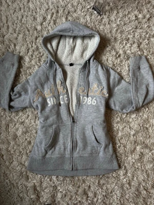 Grå hoodie med teddyfoder och tryck - Mysig grå hoodie med dragkedja och huva, fodrad med vitt teddyfoder. Framsidan har broderat texttryck 'Authentic Since 1986' i beige och vitt. Två fickor framtill och ribbade muddar vid ärmar och nederkant. Perfekt för chill dagar.