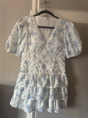 Blommig klänning - Supersöt vit kortklänning med blått blommönster, puffärmar och v-ringning. Klänningen har volanger nertill och fina spetsdetaljer vid midjan💞