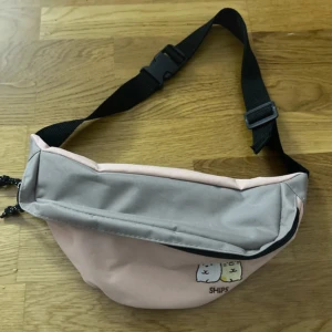 Rosa och grå magväska med SHIPS-tryck - Söt magväska i rosa och grått med justerbart svart band. Framsidan har ett tryck med två tecknade djur och texten SHIPS. Väskan har dragkedja och är tillverkad i slitstark textil, perfekt för att bära småsaker.