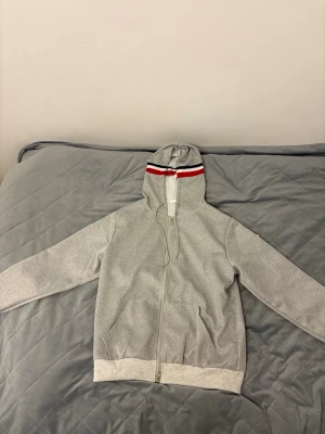 Grå hoodie med röd och blå detalj - Snygg grå hoodie från Moncler med dragkedja, huva och ribbade muddar. Huva och ärmslut har röd, vit och blå randig detalj. Klassisk logga på ärmen. Perfekt för en sportig och stilren look.