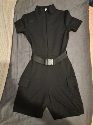 Svart kortärmad jumpsuit med bälte - Stilren svart jumpsuit med korta ärmar och shortsben. Har en hög krage, två fickor på benen och ett brett, justerbart bälte med spänne i midjan. Perfekt för en trendig och avslappnad look.