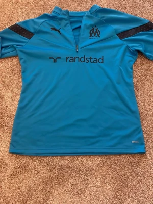 Blå träningströja OM Puma - Säljer en blå träningströja från Puma med Olympique de Marseille-logga och Randstad-tryck på bröstet. Tröjan har halv dragkedja, svarta detaljer på axlarna och är tillverkad i funktionsmaterial. Perfekt för fotbollsträning eller gym.