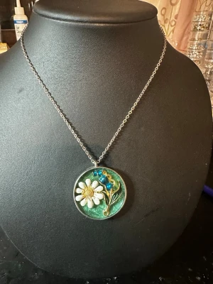 Halsband med blomma och pärldetaljer - Unikt halsband med en rund berlock i grönt, dekorerad med en vit och gul blomma samt blå pärlor och guldfärgade detaljer. Kedjan är i silverfärg och berlocken har en konstnärlig, handgjord känsla. Perfekt för dig som gillar naturinspirerade smycken och vill sticka ut.