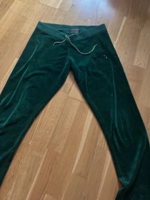 Mörkgröna velourbyxor Juicy Couture - Mörkgröna mjukisbyxor i mjuk velour från Juicy Couture. Byxorna har dragsko i midjan och raka ben. Klassisk logga broderad på ena sidan framtill. Perfekta för en chill och avslappnad stil.