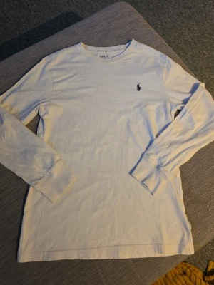 Vit långärmad tröja från Polo Ralph Lauren - Vit långärmad topp från Polo Ralph Lauren med klassisk rund hals och broderad logga på bröstet. Tillverkad i mjuk bomull som känns skön mot huden. Perfekt för en clean och stilren look.