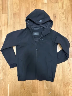 Svart vindjacka från Sail Racing - Svart vindjacka från Sail Racing med huva och dragkedja framtill. Jackan har två sidofickor, justerbar nederkant och diskret logga på bröstet. Tillverkad i ett slitstarkt syntetmaterial med mjukt foder på insidan. Perfekt för blåsiga dagar.