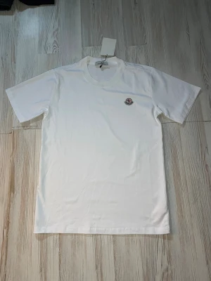 Vit Moncler t-shirt med logga - Vit t-shirt från Moncler med klassisk rund hals och korta ärmar. På bröstet sitter Monclers ikoniska broderade logga. Enkel och stilren design i mjuk bomull, perfekt för en clean och trendig look.