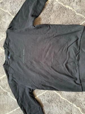 Svart sweatshirt från Robbie Moor - En stilren svart sweatshirt från Robbie Moor med diskret logga tryckt på bröstet. Tröjan har rund halsringning, ribbade muddar och är tillverkad i mjuk bomull. Perfekt för en avslappnad och trendig look. Har dock några fläckar på högra arm men det ses inte när man har på sig den.