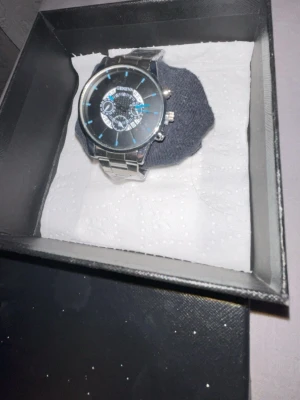 Silverfärgad klocka från Geneva - Stilren klocka från Geneva med svart urtavla och blå detaljer. Klockan har ett klassiskt runt ur och länkarmband i silverfärgad metall. Levereras i en snygg presentask, perfekt för dig som vill ha en modern accessoar.