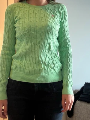 Mintgrön kabelstickad tröja Morris - Fräsch mintgrön kabelstickad tröja från Morris Lady i 100% bomull. Tröjan har rund halsringning, ribbade muddar och en liten rosa broderad logga på bröstet. Perfekt för dig som gillar stilrena och färgglada plagg.