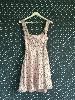 Blommig kort klänning med rosett - Supersöt ljusrosa kort klänning med blommigt mönster och lite bredare axelband. Klänningen har en fin rosett och öppen rygg.