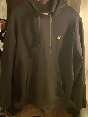 Svart hoodie från Lyle & Scott - Svart hoodie från Lyle & Scott med klassisk gul logga på bröstet. Hoodien har dragsko i huvan och en stor magficka. Perfekt för dig som gillar en stilren och enkel look. Materialet är mjukt och skönt, passar till det mesta.
