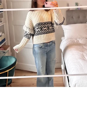 Beige stickad tröja med svart mönster - Säljer en chunky beige stickad tröja med svart grafiskt mönster och fransdetaljer på ärmarna. Tröjan har bred ribbstickning, rund hals och avslappnad passform. Perfekt för dig som gillar en trendig och mysig look.