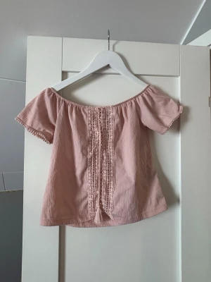 Ljusrosa offshoulder blus med spets - Söt ljusrosa offshoulder blus med korta ärmar och dekorativa spetsdetaljer framtill. Blusen har knytband med tofsar och är tillverkad i ett lätt, krinklat bomullstyg. Ena snöret har tappat tofsen, inget man tänker på däremot. Perfekt för varma dagar och ger en romantisk vibe.
