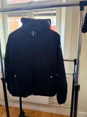 Blå  vindjacka från Moncler - Snygg svart vindjacka från Moncler med huva och dragkedja framtill. Jackan har två fickor med tryckknappar och Moncler-logga på ärmen. Perfekt för blåsiga dagar och enkel att matcha med olika outfits.