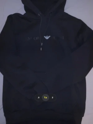 Svart hoodie från Emporio Armani EA7 - Snygg svart hoodie från Emporio Armani EA7 med logga och text framtill. Hoodien har dragsko i huvan och ribbade muddar vid ärmslut och nederkant. Perfekt för en stilren och trendig look.