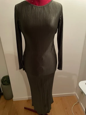 Mörkgrön långklänning med plissering - Snygg långklänning i mörkgrönt med plisserad struktur och lång ärm. Klänningen har en rund halsringning och figurnära passform som ger en stilren och modern look. Perfekt för dig som gillar minimalistisk stil.