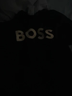 Svart t-shirt med BOSS-tryck - Svart t-shirt från BOSS med stort vitt logotryck på bröstet. Klassisk passform och rund halsringning, perfekt för en clean och stilren look.