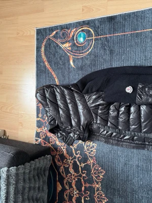 Svart dunväst från Moncler - Snygg svart dunväst från Moncler med glansig quiltad framsida och stickad rygg. Västen har en hög krage och Moncler-logga på bröstet. Perfekt för lager-på-lager och ger en cool vibe till din outfit.