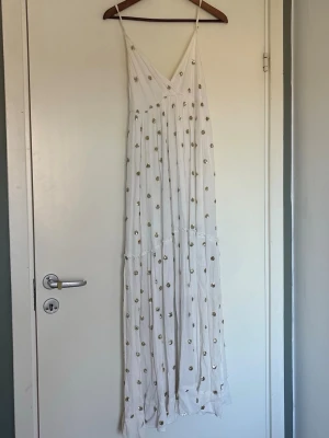 Vit långklänning med guldprickar - Säljer en vit långklänning med tunna axelband och v-ringning. Klänningen har ett flowy fall och är dekorerad med glittriga guldprickar över hela tyget. Den har tappat en av ”bollarna” i knytningen i ryggen 