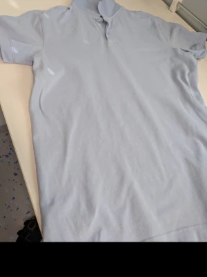 Ljusblå pikétröja från No 15 - Snygg ljusblå pikétröja från No 15 med klassisk krage och två knappar. Tillverkad i 100% bomull för en skön och luftig känsla. Perfekt till jeans eller shorts för en clean och enkel stil.