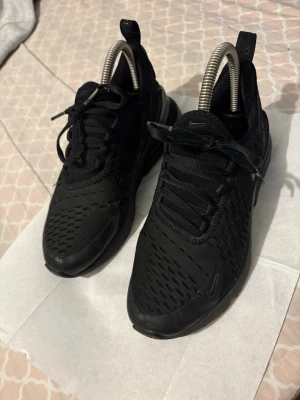 Svarta Nike Air Max 270 sneakers - Snygga helsvarta Nike Air Max 270 sneakers med mesh-överdel och synlig Air-enhet i hälen. Skorna har en sportig siluett, klassiska snörningar och diskreta Nike-loggor på sidan och tungan. Perfekta för dig som gillar streetwear och bekväma skor.