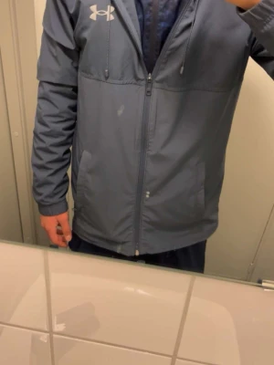 Blå vindjacka från Under Armour - Snygg blå vindjacka från Under Armour med huva och dragkedja framtill. Jackan har två sidofickor och logga på bröstet samt stor logotyp på ryggen. Tillverkad i lätt och tåligt material. Pris kan diskuteras!!