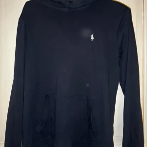 Mörkblå hoodie från Polo Ralph Lauren - Mörkblå hoodie från Polo Ralph Lauren med klassisk vit logga på bröstet. Hoodien har känguruficka framtill och långa ärmar. Perfekt för en avslappnad och stilren look.