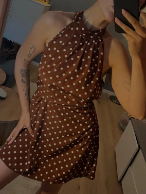 Brun prickig halterneckklänning - Säljer en brun klänning med vita prickar och halterneck. Klänningen har en markerad midja med knytband och kort kjol. Man kan knyta bak eller fram. Tyget är lätt och faller snyggt. Vintage från märket Love21 storlek S 