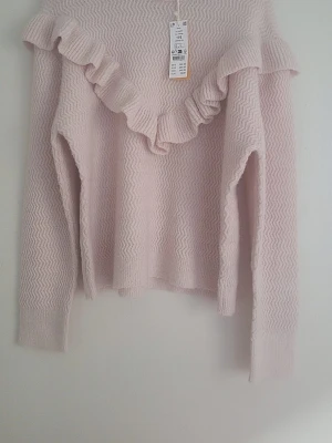 Ljusrosa stickad tröja med volang - Supersöt ljusrosa stickad tröja med volangdetalj över bröstet och axlarna. Tröjan har ett diskret sicksackmönster i materialet och långa ärmar. Perfekt för dig som vill ha en mjuk och trendig look. Helt ny och oanvänd. Prislapp finns kvar!