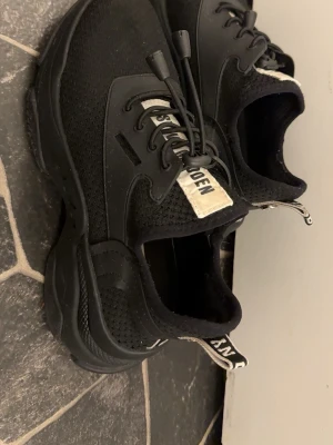 Svarta sneakers från Stadsmissionen - Svarta sneakers med chunky sula och stickad mesh-överdel. Skorna har svarta snören med snabblås och vit etikett med text på plösen och hälen. Snygg och sportig design som passar till streetwear. Materialet är en mix av textil och syntet.
