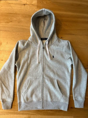 Grå hoodie från Polo Ralph Lauren - Snygg ljusgrå hoodie med dragkedja från Polo Ralph Lauren. Klassisk design med vit dragsko i huvan, två fickor framtill och broderad logga på bröstet. Mjuk bomullsblandning som är skön att ha på sig.