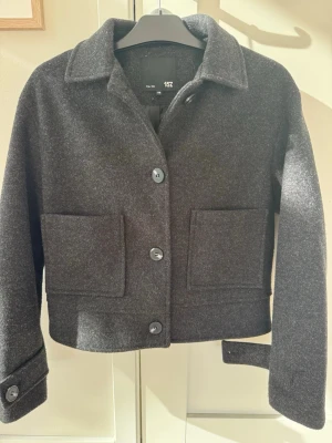 Mörkgrå ullcardigan från 157 - Perfekt vår/höst jacka. Stilren mörkgrå cardiganjacka från 157 i ullmix. Jackan har stora framfickor, klassisk krage och knäppning med fyra knappar framtill. Ärmarna har dekorativa knappar vid mudden och modellen är något boxig och croppad. Perfekt för lager på lager under kyliga dagar. Har dock tappat en knapp på ena ärmen men den går säkert att sy dit enkelt.