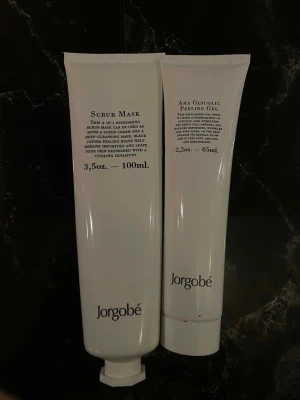 Jorgobé Scrub Mask & Peeling Gel - Säljer två vita tuber från Jorgobé: en Scrub Mask (100 ml) och en AHA Glycolic Peeling Gel (65 ml). Båda produkterna är perfekta för ansiktsvård och exfoliering. Tubernas design är stilren och minimalistisk med svart text på vit bakgrund.