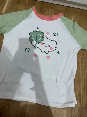 Söt t-shirt med katt och fyrklöver - T-shirt med mintgröna ärmar och rosa krage. Framsidan har ett gulligt tryck av en katt med fyrklöver och rosa detaljer. Perfekt för dig som gillar kawaii-stil och vill sticka ut lite extra.