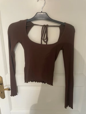 Brun långärmad topp med öppen rygg från Nelly  - Snygg brun långärmad topp med öppen rygg och knytband i nacken. Toppen är ribbad och har en croppad passform med vågig kant nertill.