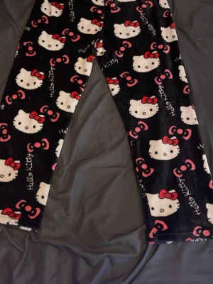 Hello Kitty pyjamasbyxor svart - Mysiga pyjamasbyxor i svart med Hello Kitty-motiv och röda rosetter. Byxorna är i mjukt fleecematerial och har ett lekfullt mönster med vita Hello Kitty-ansikten och text. Perfekta för chillkvällar hemma.