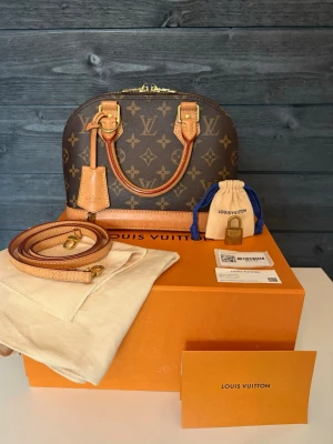 Louis Vuitton Alma BB Monogram i mycket fint skick säljes - Väskan är köpt 2022 och är en nyare modell utrustad med Louis Vuittons inbyggda microchip (NFC-chip) istället för datumkod.  Sparsamt använd och väl omhändertagen. Insidan är ren och fin, dragkedja fungerar perfekt och canvasen är utan skador. Lädret har utvecklat en naturlig patina.                                                                              Medföljer: • Originalkartong • Dustbag • Axelrem • Hänglås & nycklar • Louis Vuitton-kort • Bild på originalkvitto 