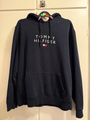 Mörkblå hoodie från Tommy Hilfiger - Mörkblå hoodie med broderad Tommy Hilfiger-logga och flagga på bröstet. Klassisk känguruficka framtill, justerbar huva med snören och ribbade muddar. Perfekt för en avslappnad och stilren look.
