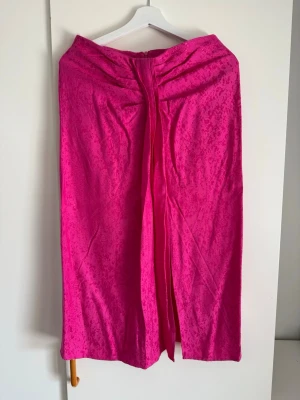 Koton fuchsia midi kjol, storlek 40 - En midi kjol i en livlig fuchsiafärg med ett diskret mönster. Kjolen har en elegant knuten detalj framtill som skapar ett draperat utseende och en slits. Dragkedjan är placerad baktill för en smidig passform.  Buy shipping label or pick it up from Farsta Centrum in Stockholm.