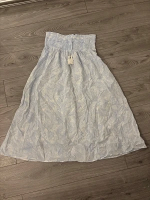 Ljusblå långkjol med snörning - Ljusblå långkjol med vitt bladmönster och bred smock i midjan. Kjolen har snörning med tofsar framtill och är gjord i ett tunt, luftigt material som passar perfekt till varmare dagar. Snygg att styla med en enkel topp eller crop top.