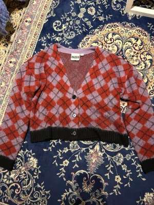 Lila och röd argylekofta Ragged Priest - Kofta från Ragged Priest i lila och rött med argylemönster. Modellen är croppad med v-ringning och svarta ribbade muddar. Tre knappar framtill och lång ärm. Perfekt för dig som gillar retrovibbar och statementplagg.