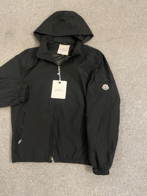 Svart vindjacka från Moncler - Svart vindjacka från Moncler med huva och dragkedja framtill. Jackan har två sidofickor med dragkedja och en ficka med knapp på ärmen där Moncler-loggan sitter. Tillverkad i lätt och slitstarkt material, perfekt för blåsiga dagar.