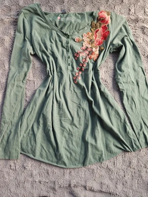 Grön blus med broderi och knappar - Superfin grön blus med långa ärmar och dekorativa knappar framtill. Snygg broderad blomdetalj i rött och rosa vid axeln som ger en unik touch. Blusen är insydd vid midjan och är tillverkad i mjukt tyg, perfekt för en trendig vardagslook!❤️ Hör gärna av er vid fler frågor