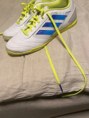 Futsal Skor Adidas - Snygga vita Adidas futsalskor med neon-gula snören och detaljer samt blå ränder på sidan. Skorna har låg profil, tålig syntetöverdel och greppvänlig yttersula för spel på konstgräs eller inomhus. Perfekta för dig som vill sticka ut på planen.