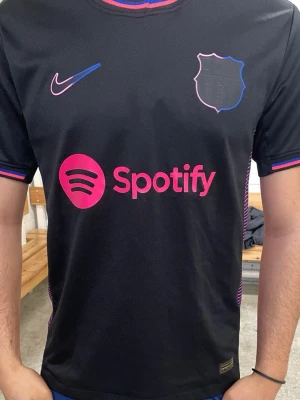 Svart FC Barcelona matchtröja Nike - Snygg svart FC Barcelona matchtröja från Nike med rosa och blå detaljer. Tröjan har korta ärmar, rund hals och tryck med Spotify-logga på bröstet. Tillverkad i lätt och ventilerande funktionsmaterial, perfekt för fotboll eller träning.